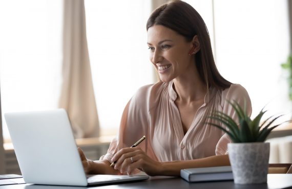 Smiling young woman wearing headphones sit at desk working at laptop making notes, happy millennial female in modern earphones study watch webinar on computer handwrite in notebook