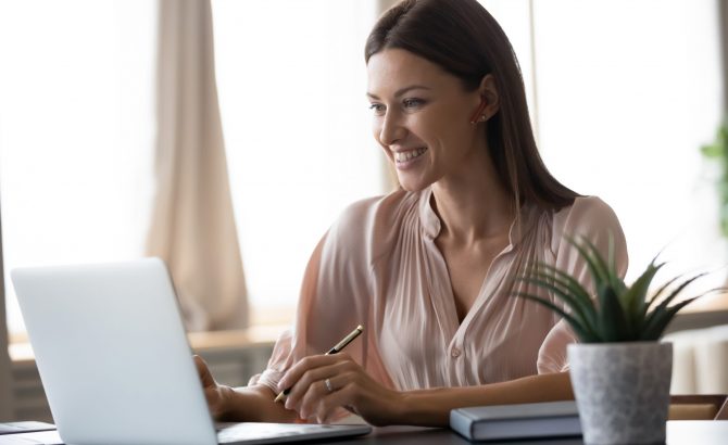 Smiling young woman wearing headphones sit at desk working at laptop making notes, happy millennial female in modern earphones study watch webinar on computer handwrite in notebook